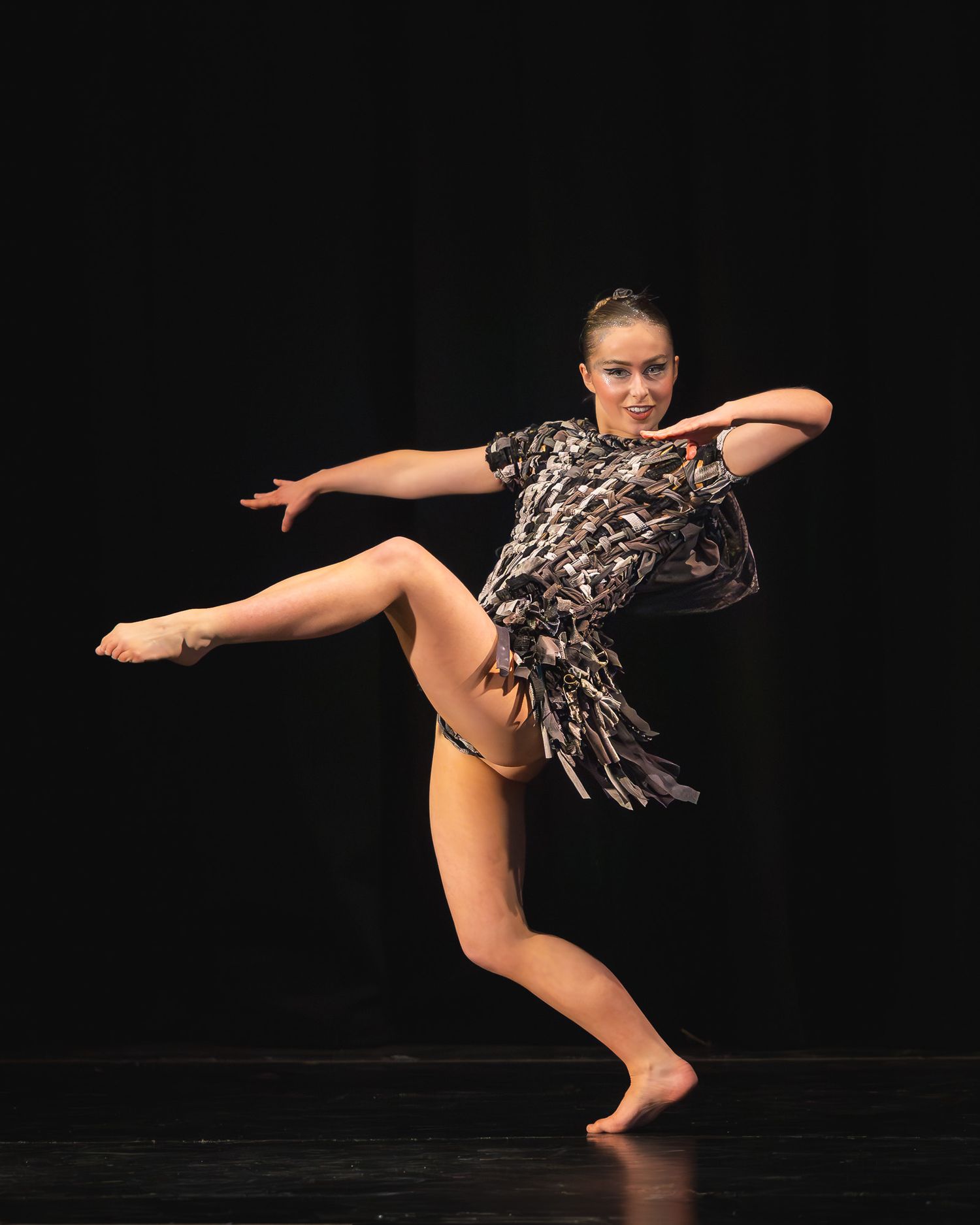 A dancer performs a dynamic solo on stage, wearing a feathered costume, balancing on one foot with leg raised.