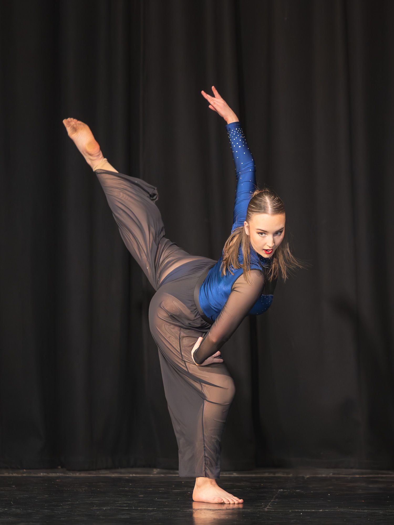 Young female dancer performs a high leg extension on stage wearing a blue sparkle top and gray pants.