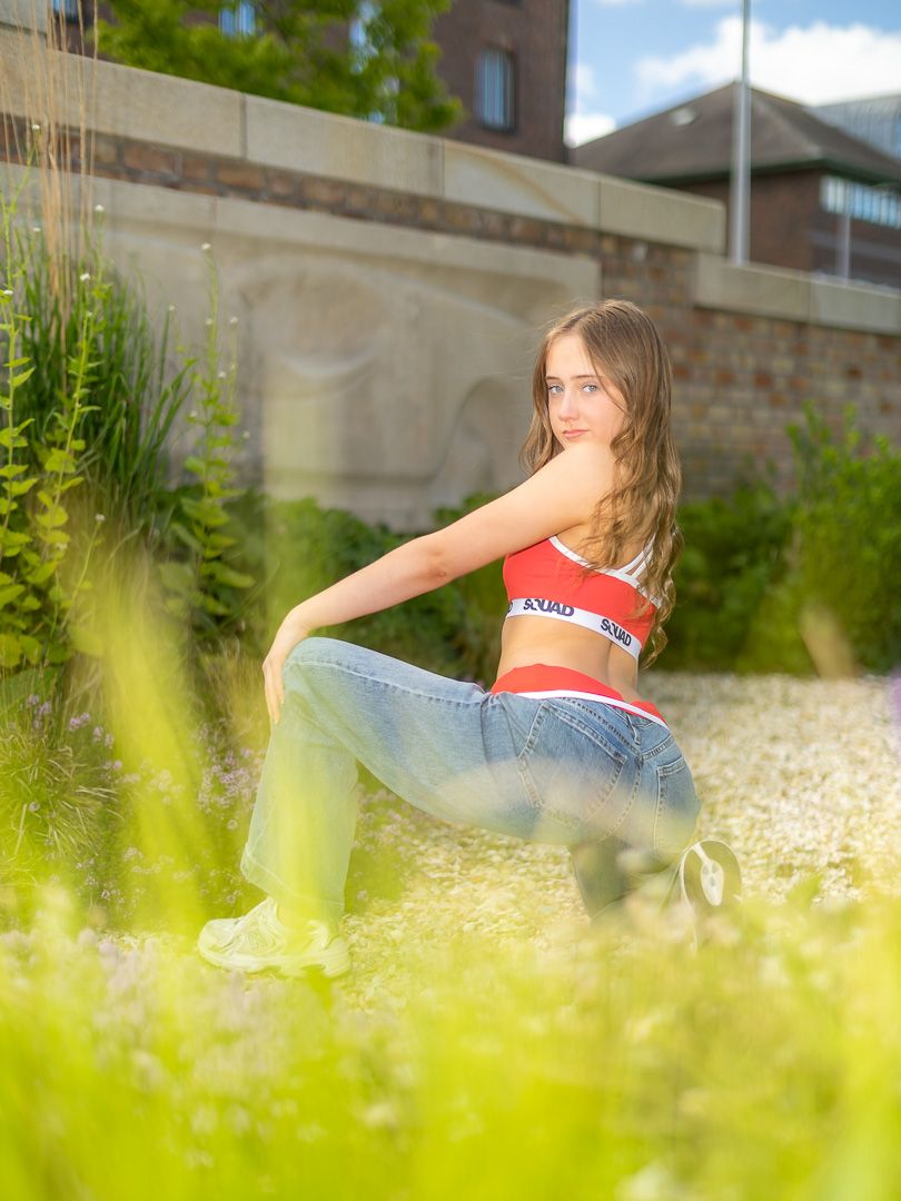 Young woman in red crop top and jeans sits gracefully among lush green plants in a sunny garden.