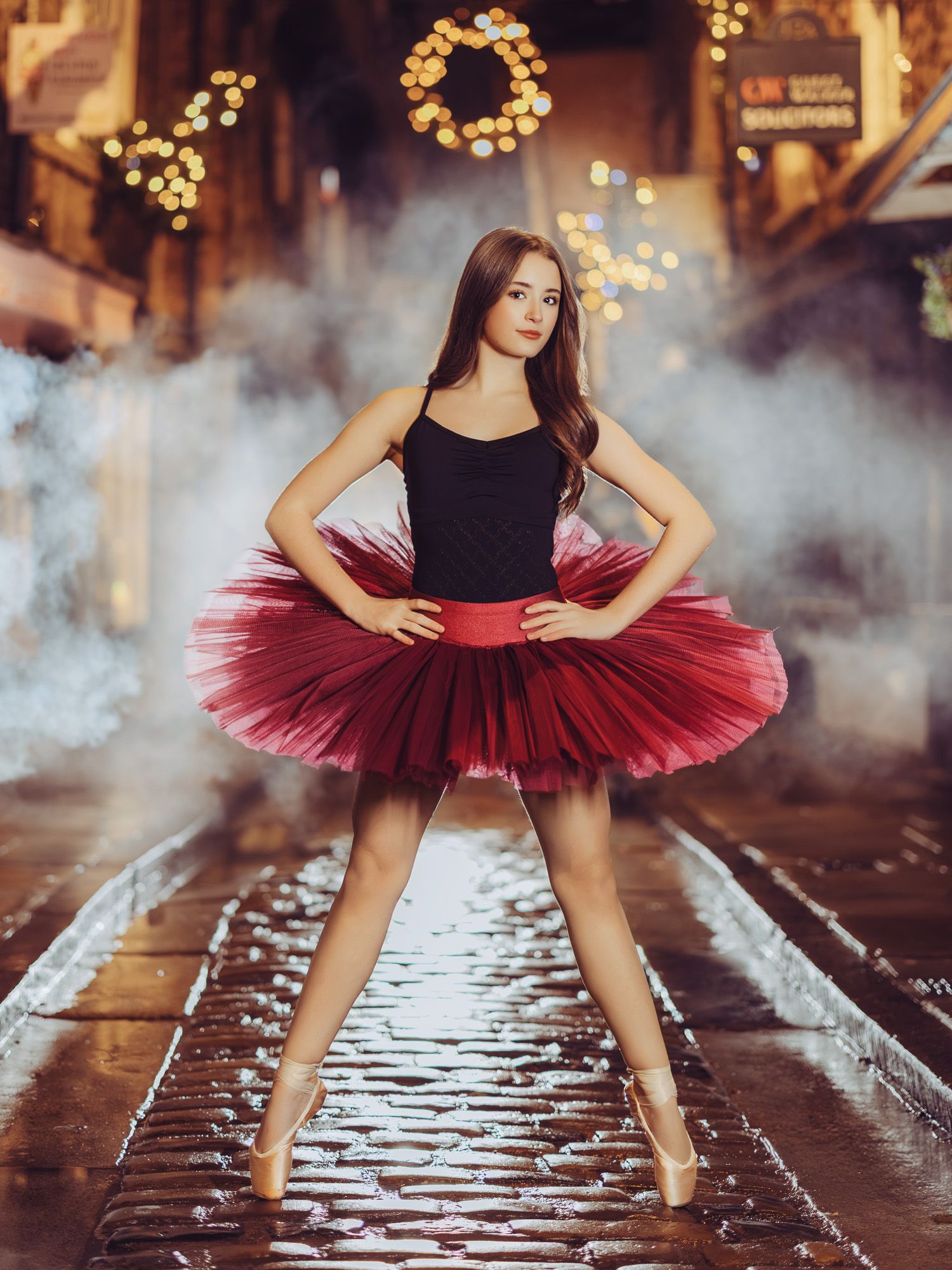 A ballerina poses in a red tutu and pointe shoes on a wet street with festive bokeh lights in the background.