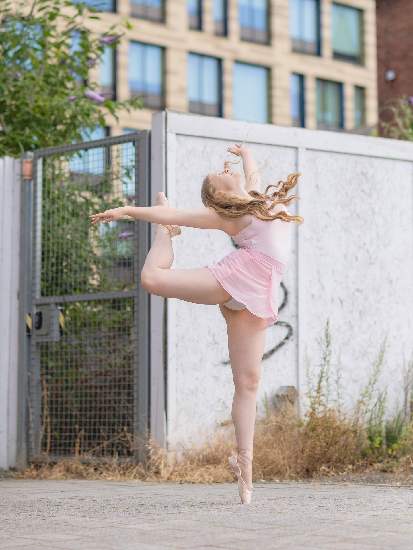 A ballet dancer in pink performs an arabesque on pointe outdoors against an urban wall.