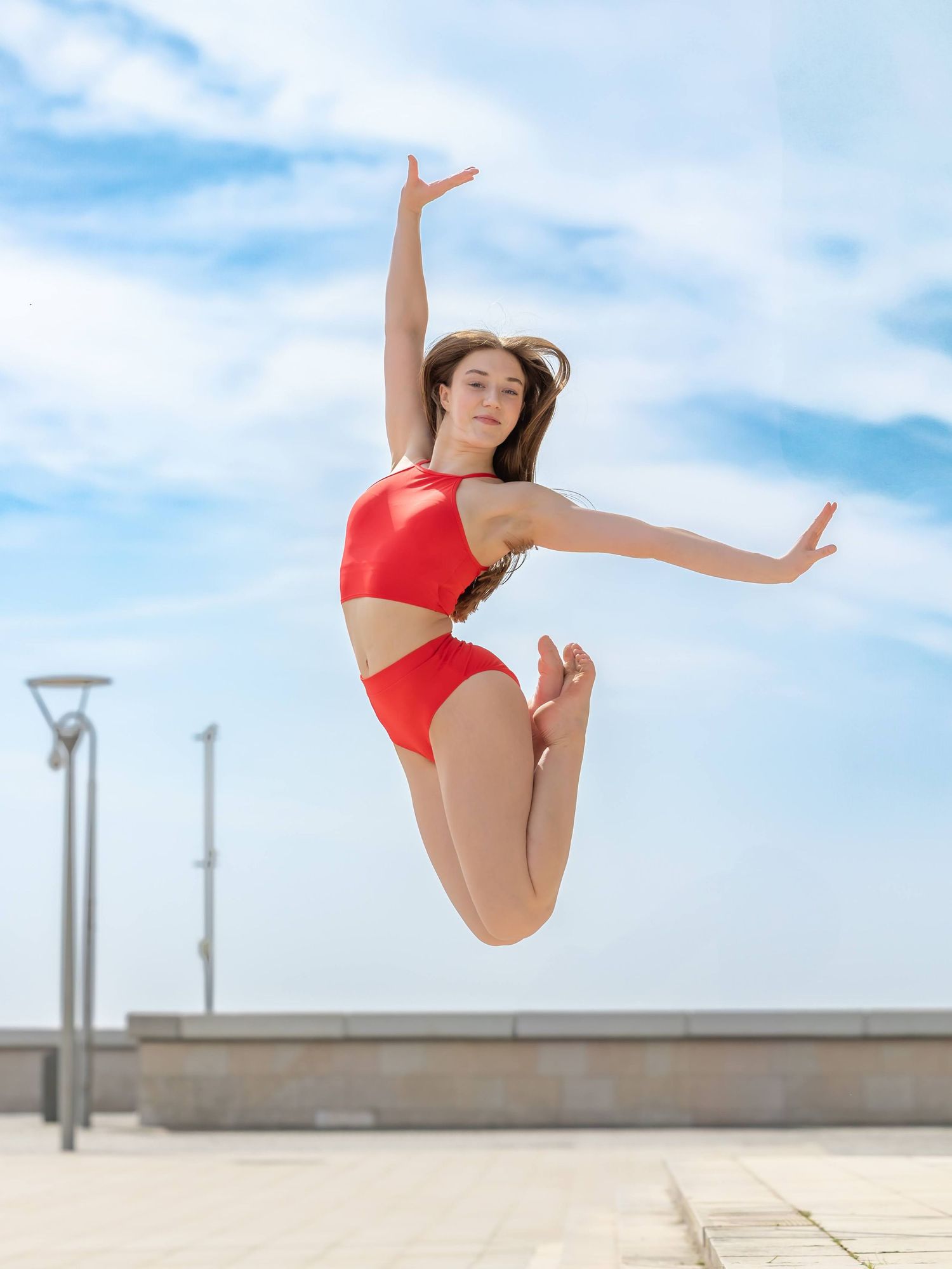 A dancer in a red leotard performs a graceful leap against a bright blue sky backdrop.