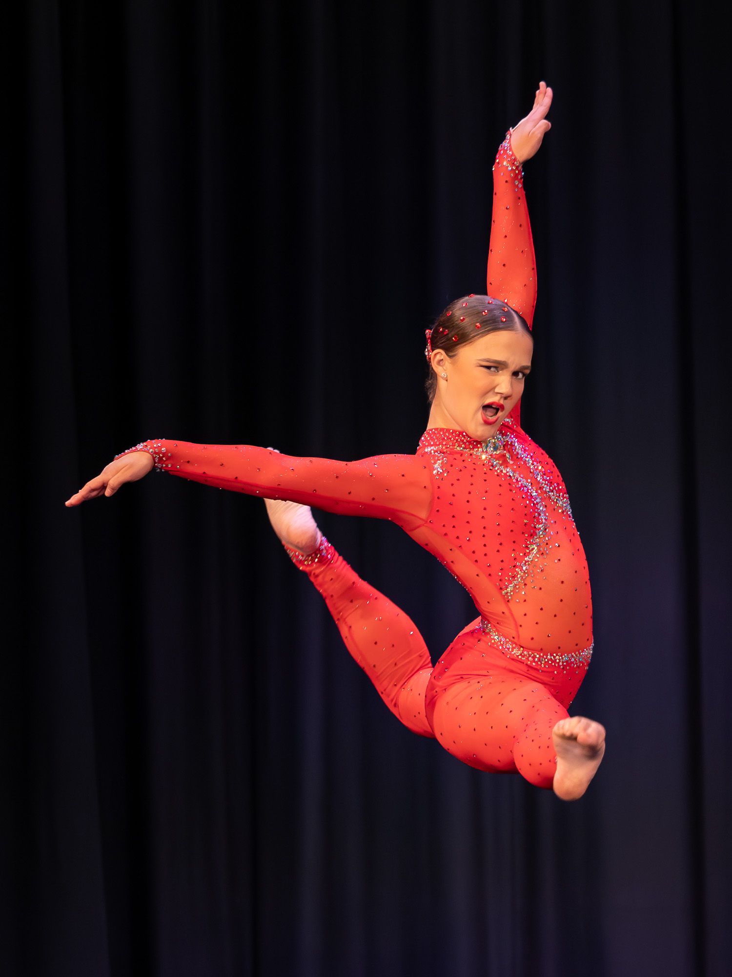 A dancer in a red rhinestone catsuit leaps mid-air during a dynamic performance on a darkened stage.