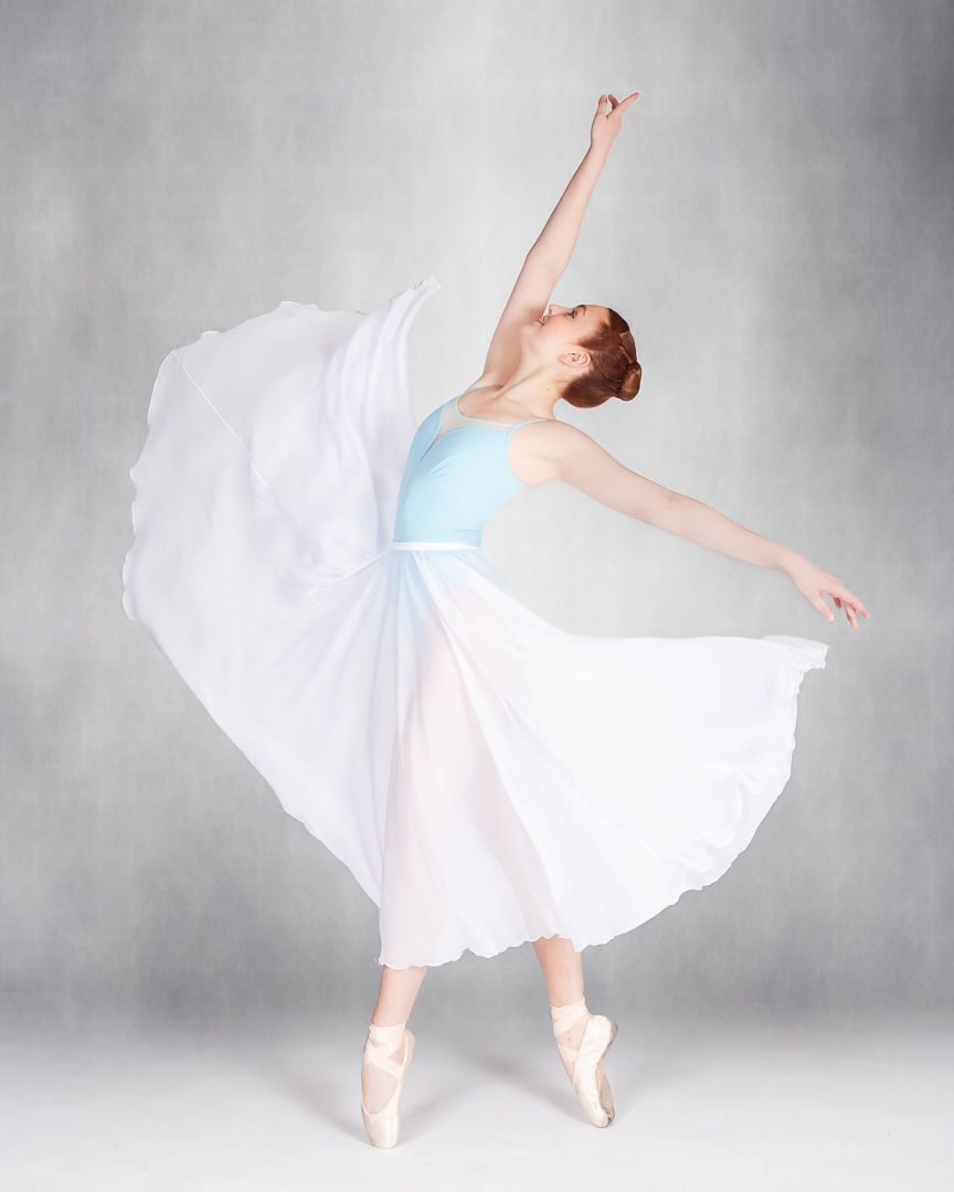 A graceful ballerina in a white flowing skirt and blue leotard performs an arabesque on pointe shoes.