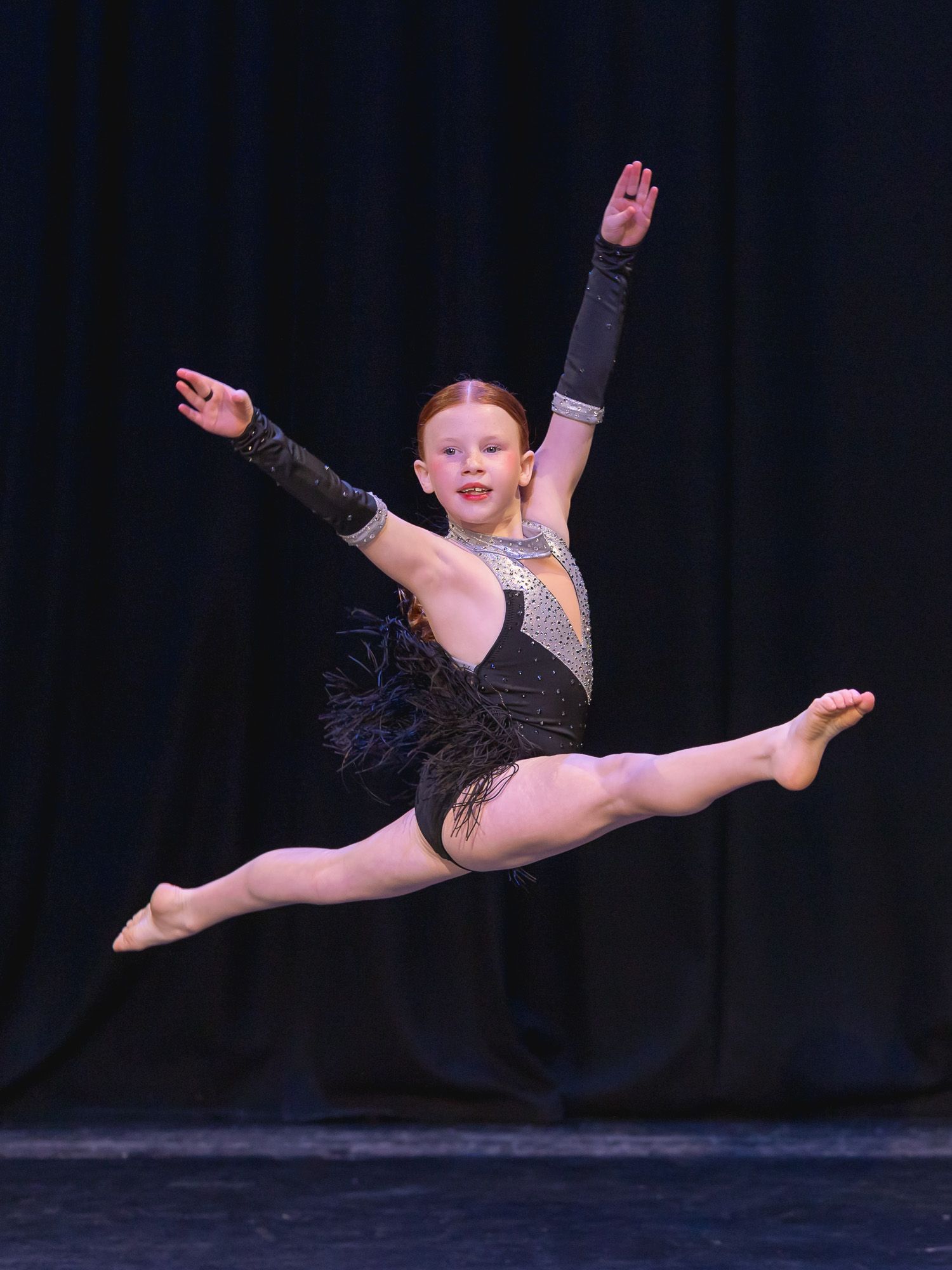 Dancer executes powerful leap in sparkly black costume against dark background.
