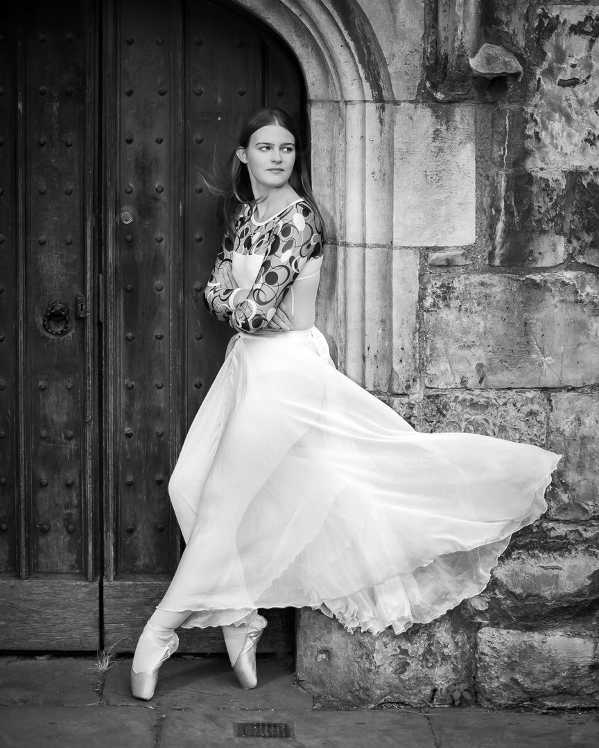 Dancer in flowing white dress performs graceful movements against stone wall backdrop.