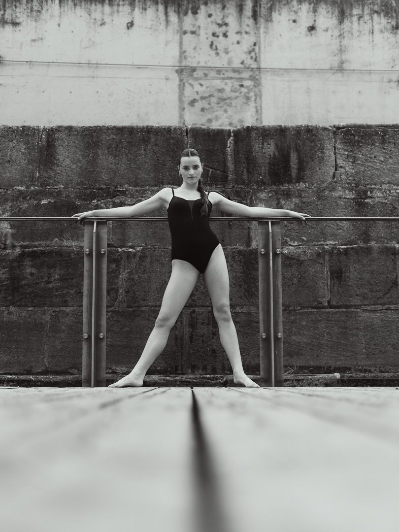 Ballet dancer in black leotard posing at outdoor barre in black and white photo.