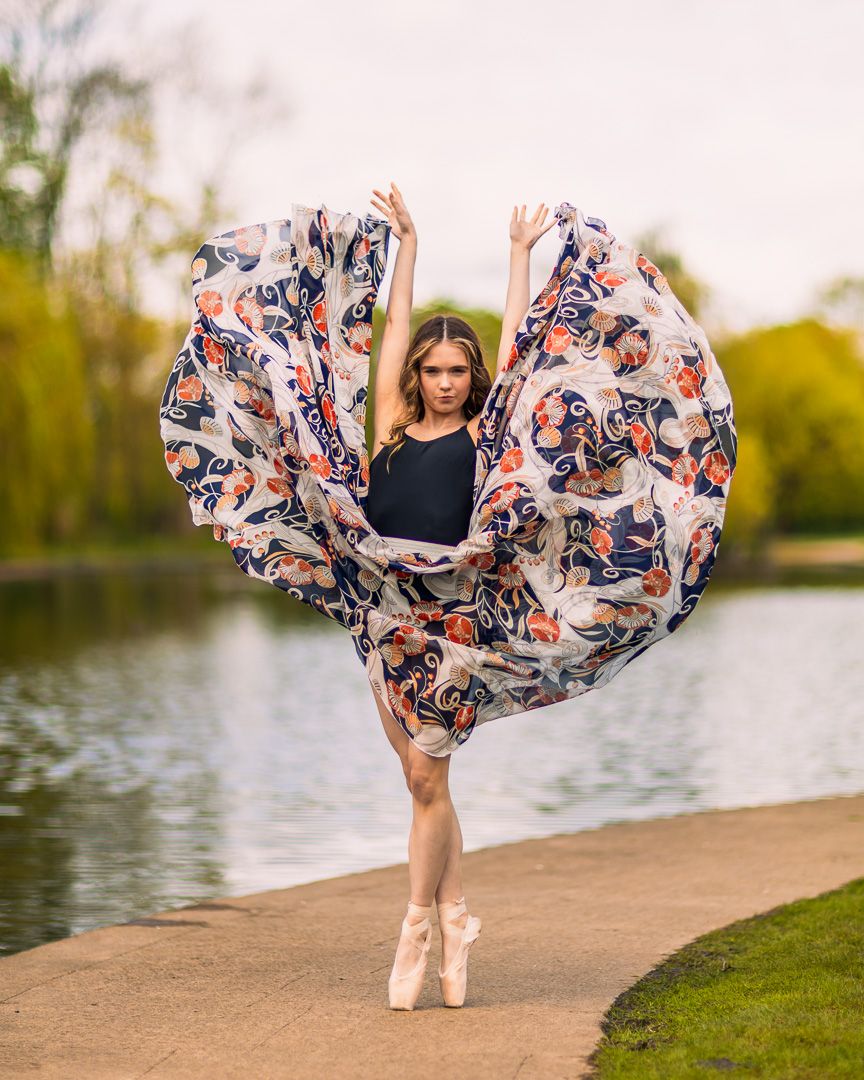 A ballerina in pointe shoes poses gracefully outdoors, her floral skirt billowing wide near a serene pond.