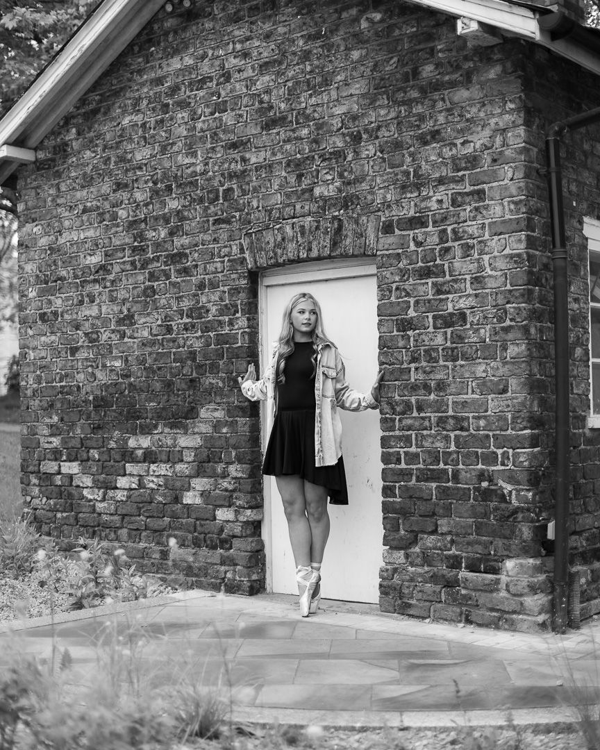Black and white photo of a blonde woman in a dress and jacket posing in a white doorway of a brick building.