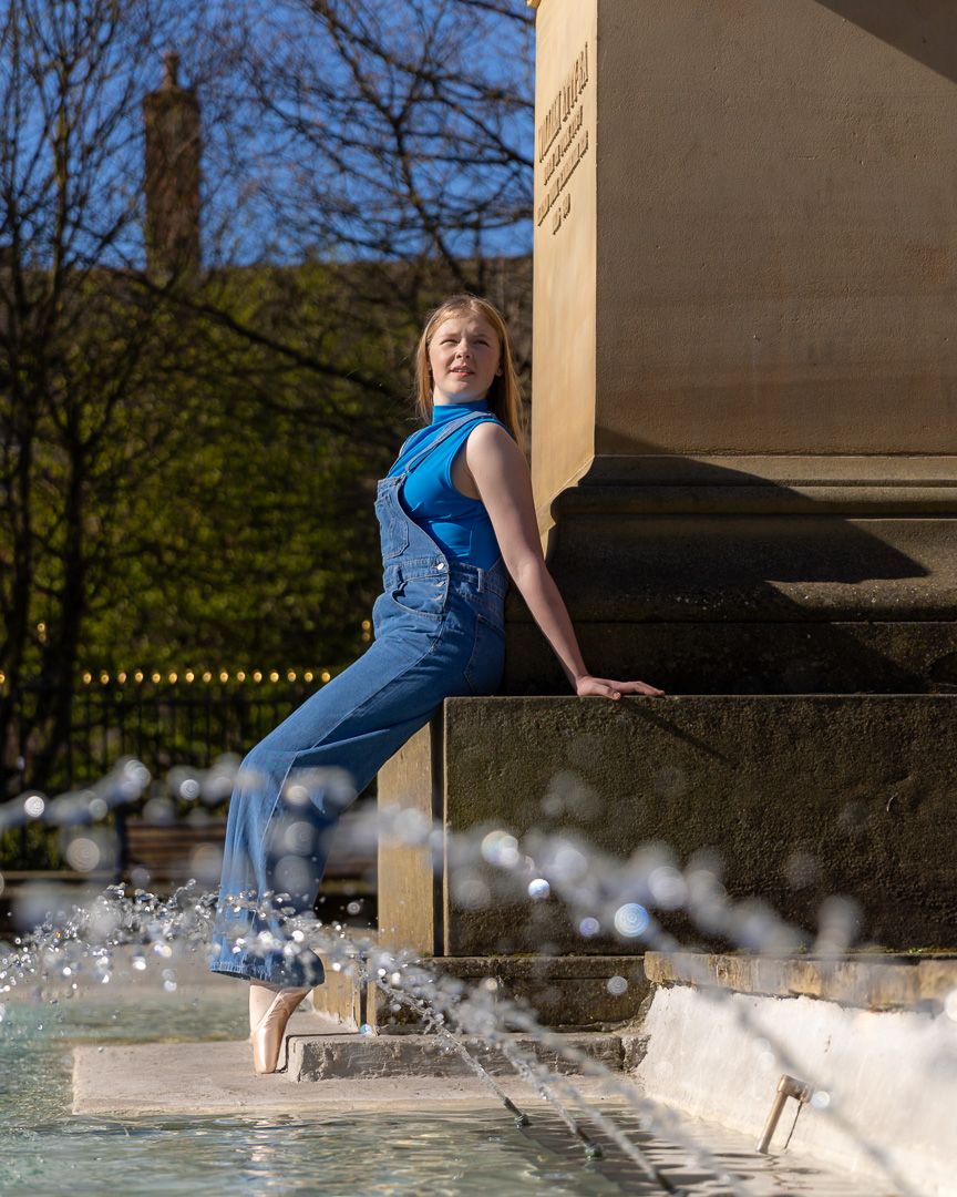 Dancer in denim overalls poses playfully by fountain with water splashing.