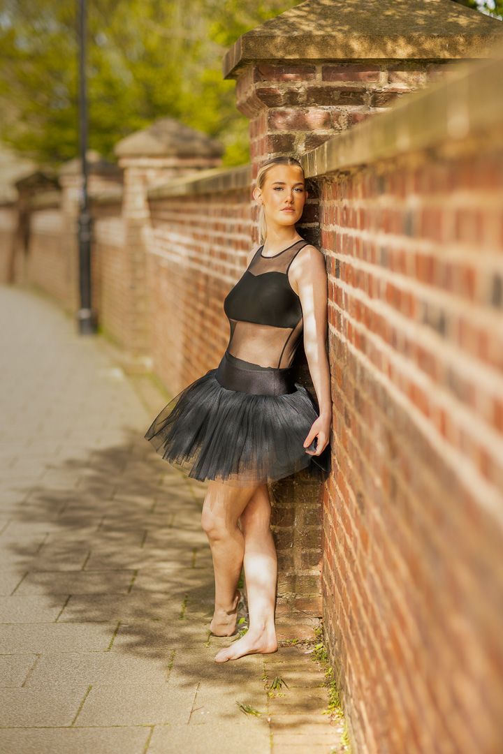 Dancer in black tutu poses gracefully against brick wall.