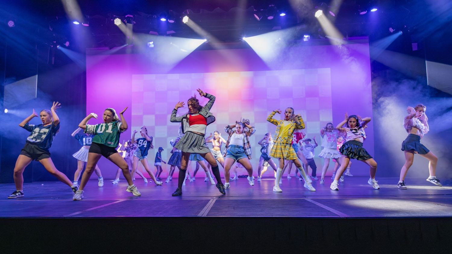 Colorful stage performance with dancers in vibrant costumes under purple and blue lights on a decorated set.