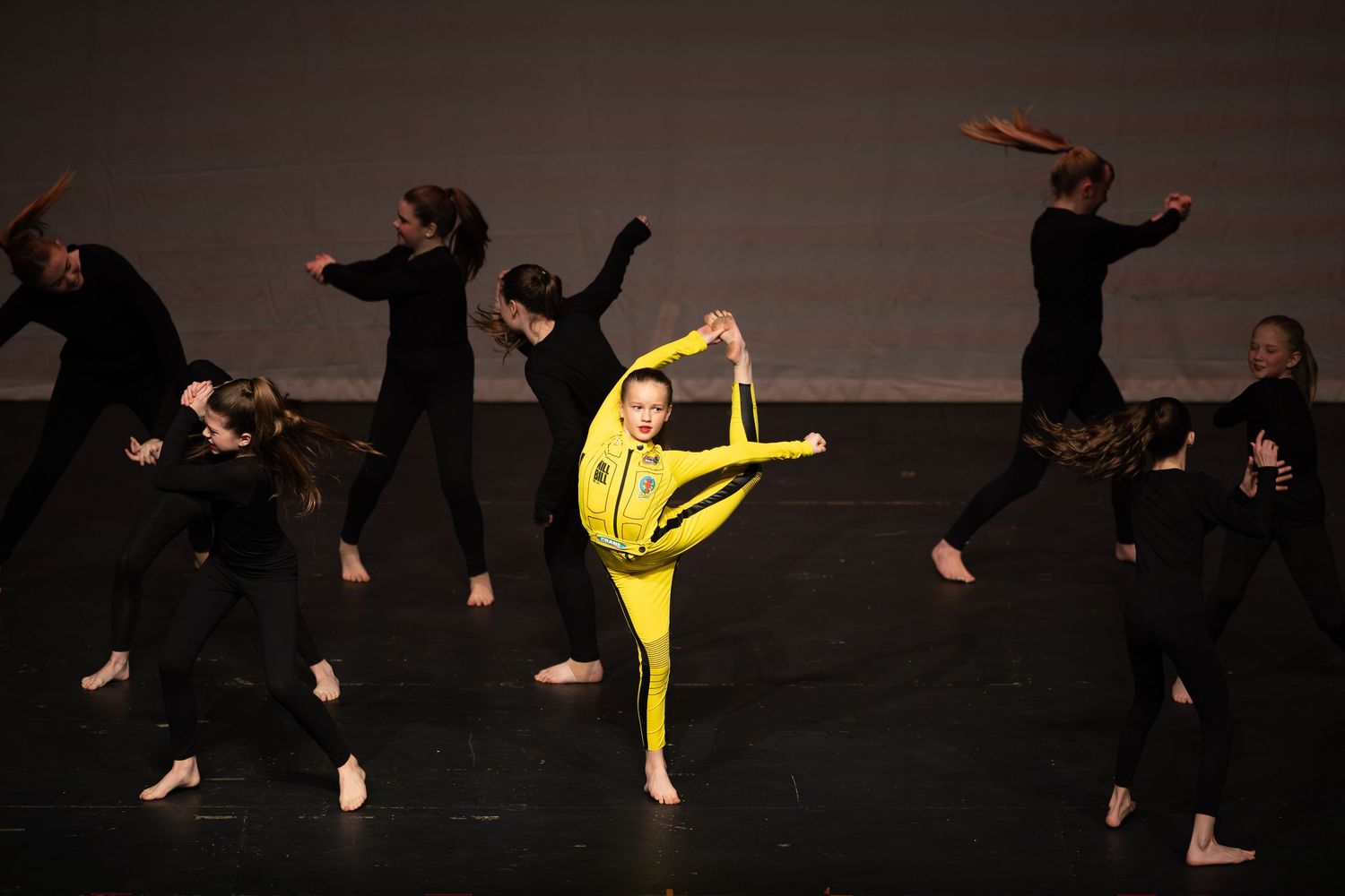 A dancer in yellow performs a high kick on stage surrounded by dancers in black during a contemporary dance performance.