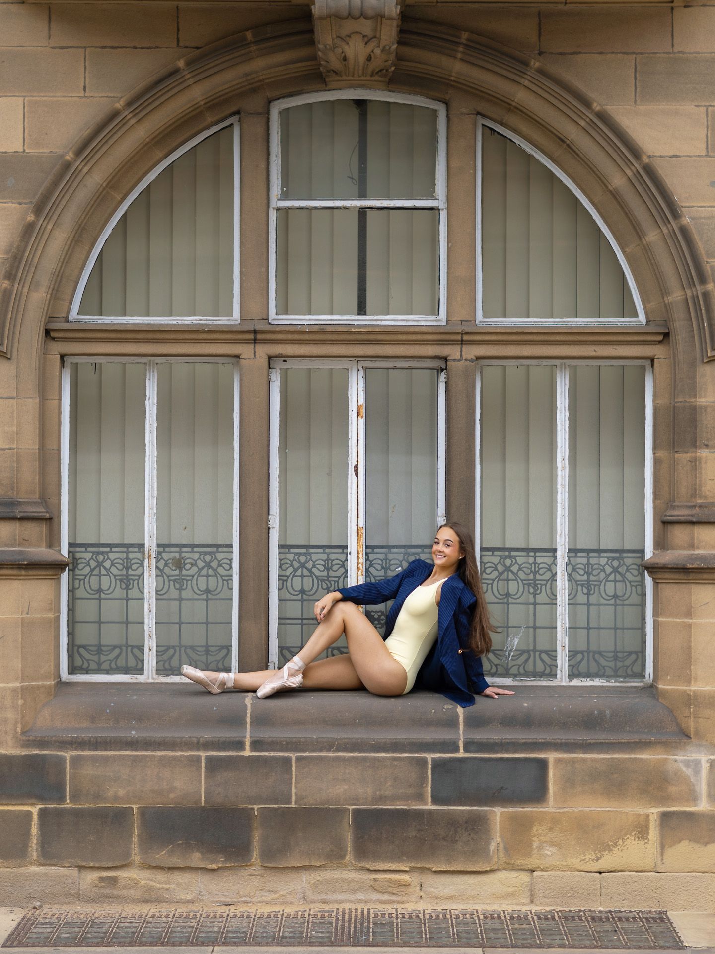 Woman in blue blazer and yellow bodysuit poses on stone window ledge of ornate arched building.
