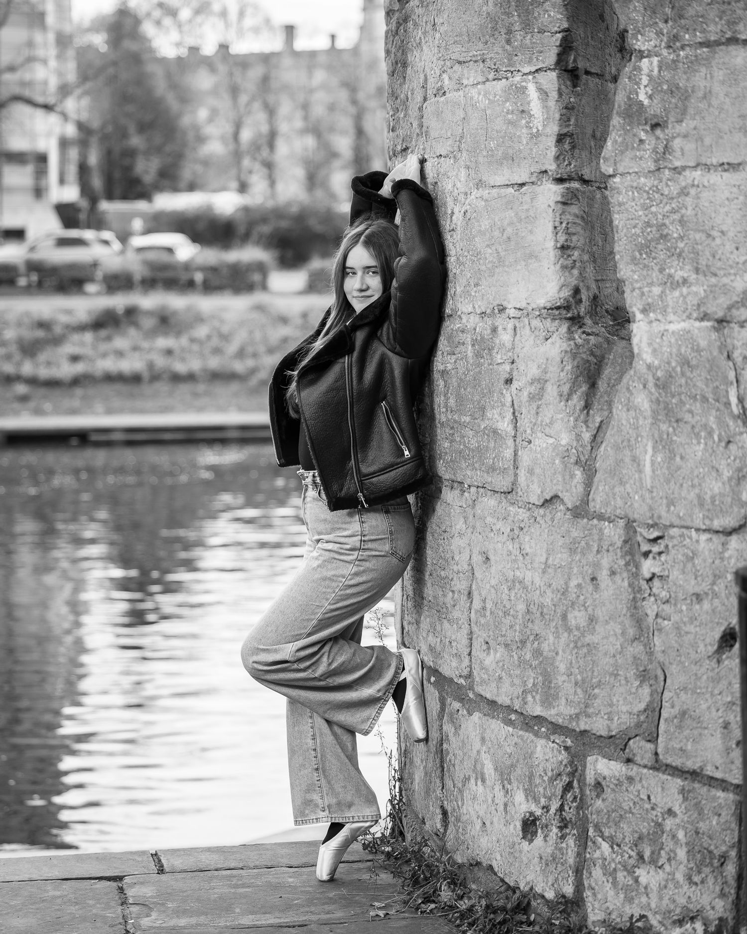 Black and white portrait of a person in a leather jacket and wide-leg pants posing against a stone wall by the water.