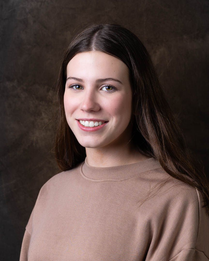 Young woman with long brown hair smiling, wearing a tan sweater, posed against a dark studio background.