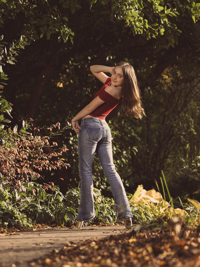Young woman in red top and blue flare jeans posing on a sunlit garden path surrounded by lush green foliage.
