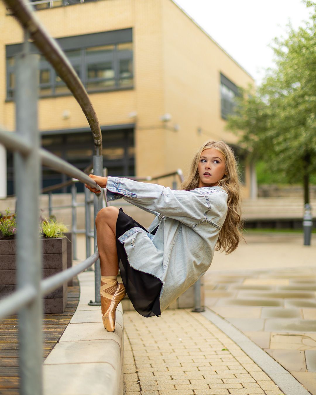 Young blonde dancer in ballet pointe shoes poses dramatically on urban steps, holding metal railing outdoors.