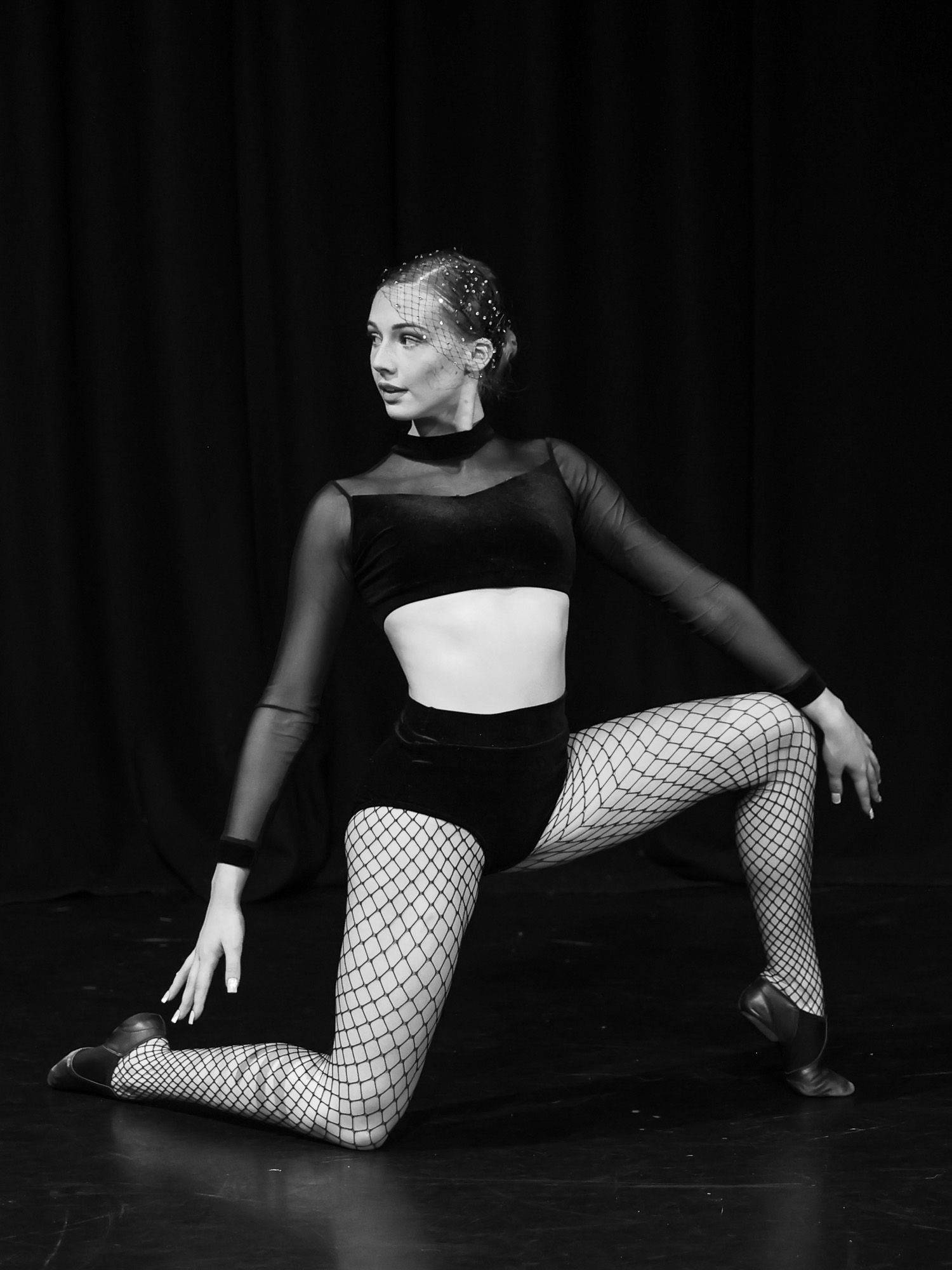 Black and white photo of a dancer in fishnet tights and velvet crop top striking a dramatic pose on stage.