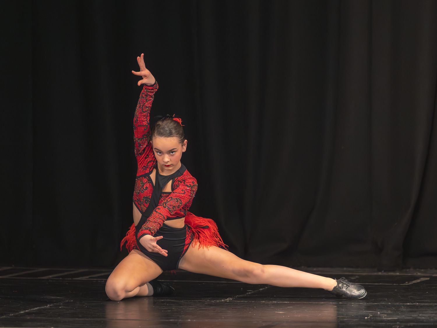 Young female dancer in red and black costume performs a dramatic split pose on stage under spotlight.