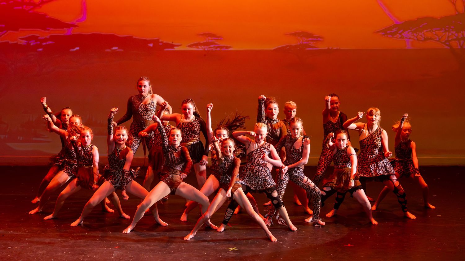 A group of dancers perform on stage under dramatic red lighting during a vibrant dance recital performance.