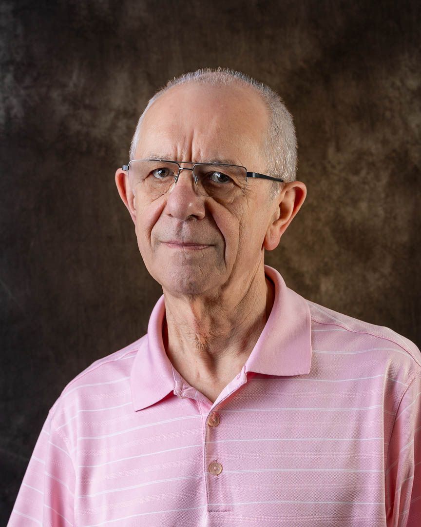Older man with glasses and short gray hair wearing a pink striped polo shirt, posing for a professional portrait.
