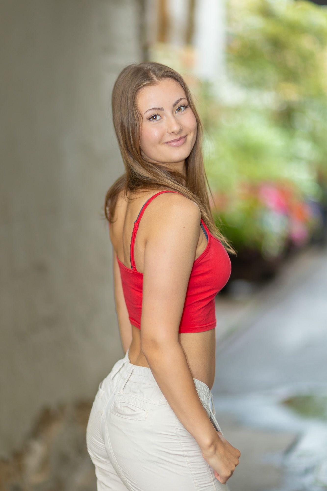 Model wearing red top and light pants poses against outdoor backdrop in casual style.