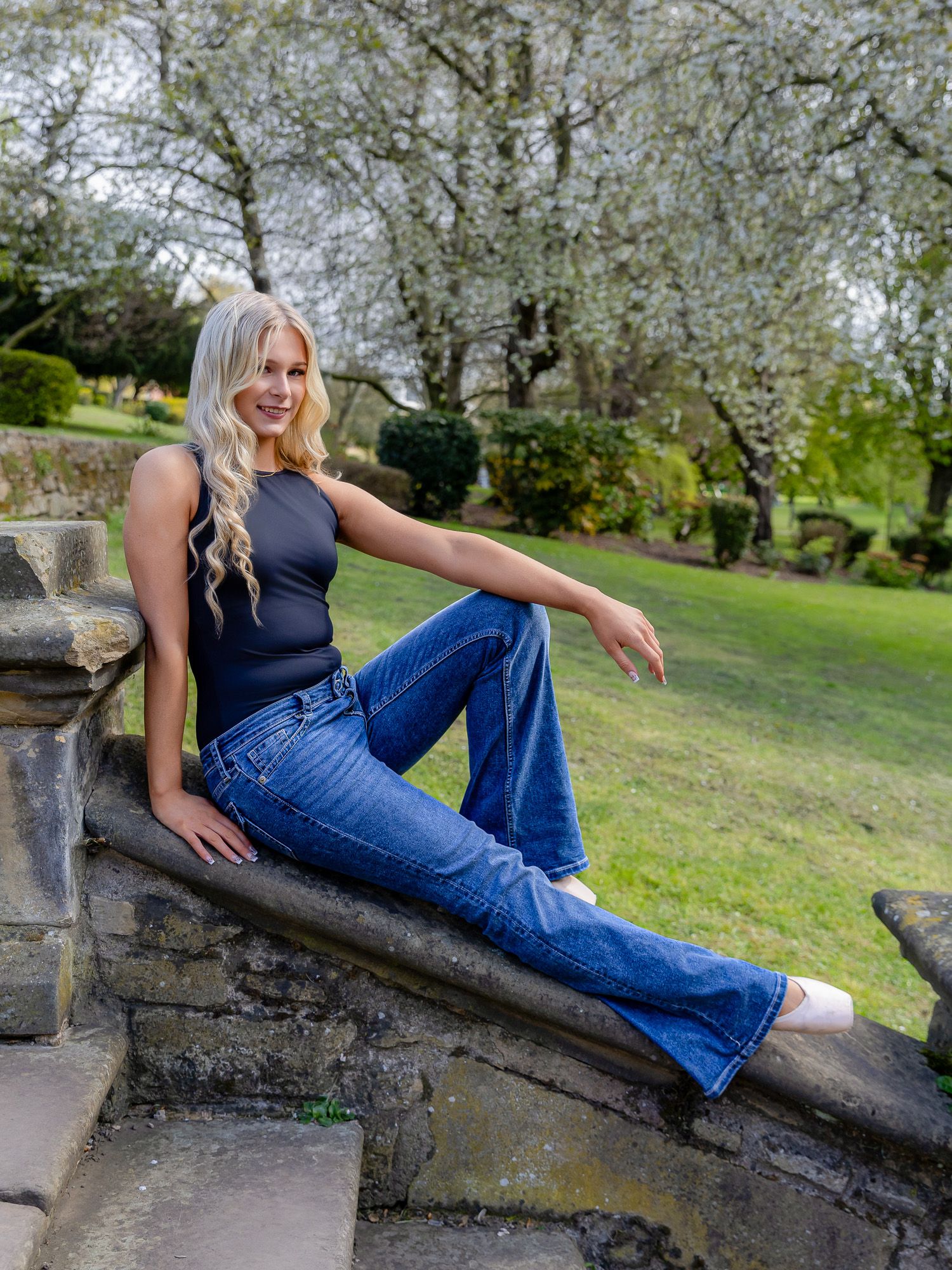 Blonde woman in dark bodysuit and flare jeans posing on stone steps in a lush green garden.