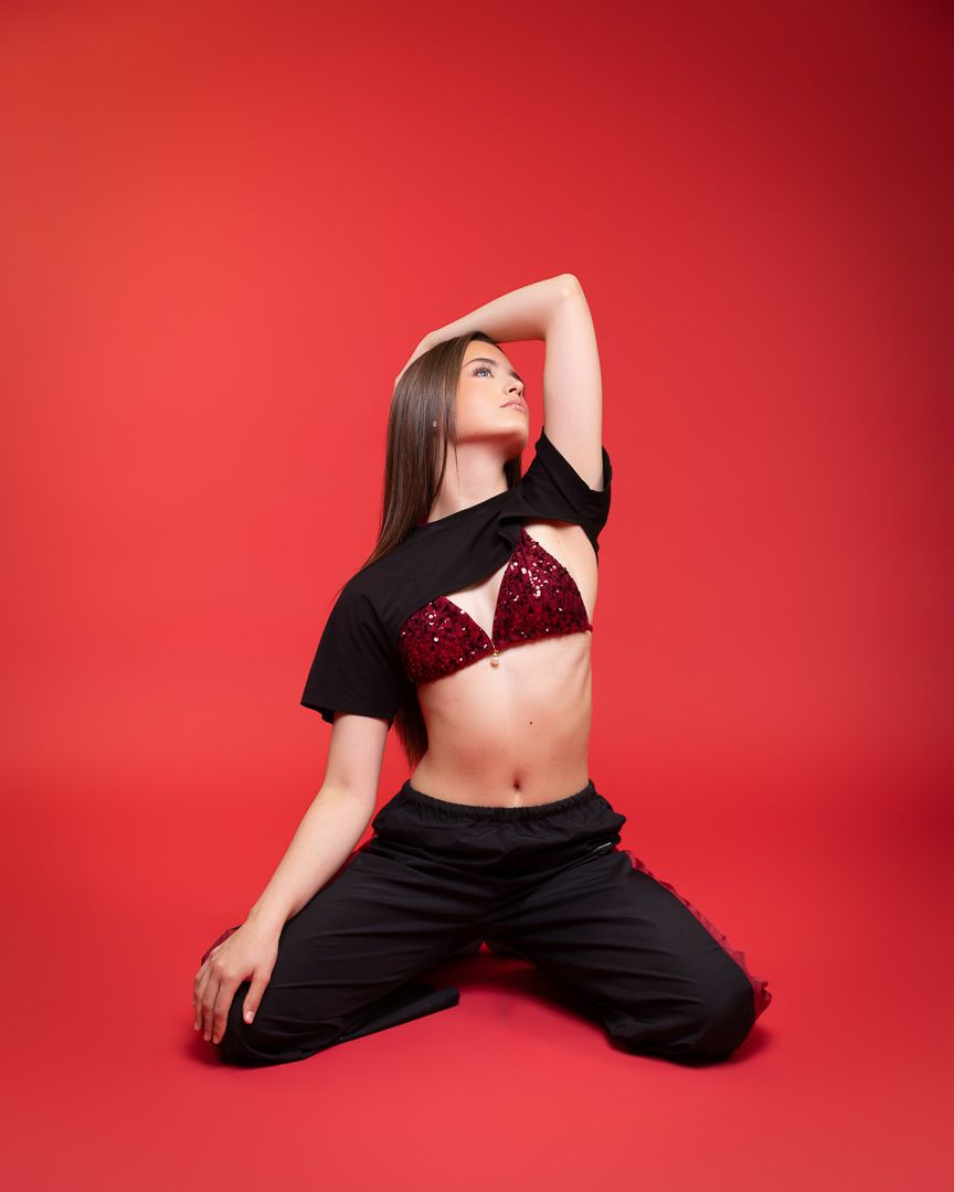 A young woman poses on her knees against a red background wearing black pants and a sparkly burgundy bralette.