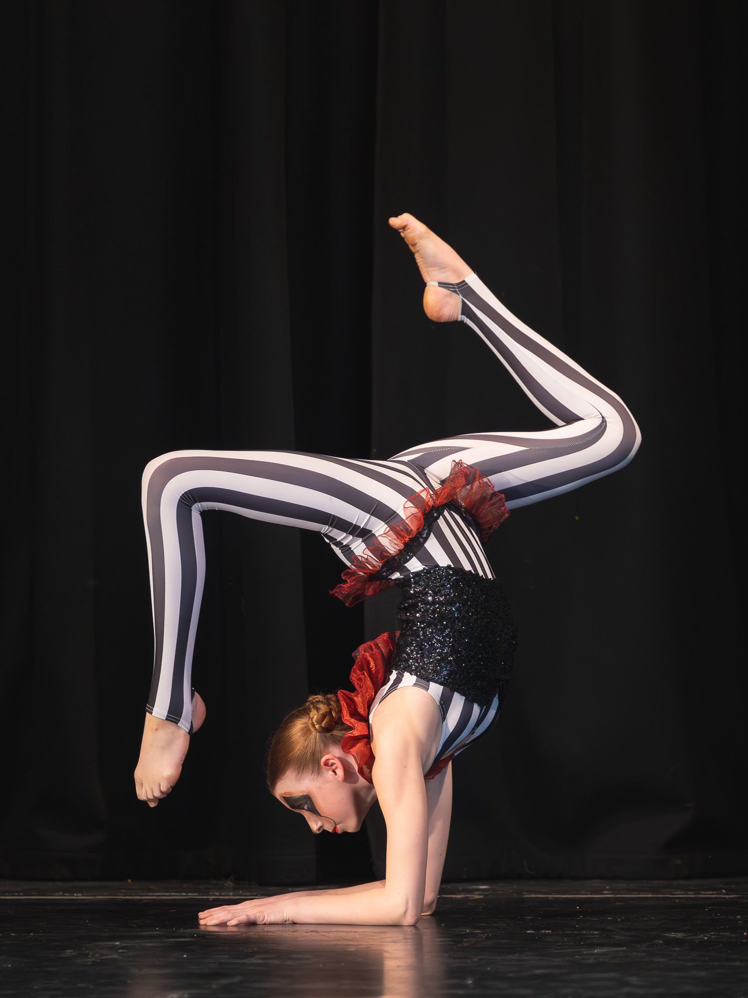 Young gymnast in striped black and white leotard performs a backbend contortion pose on stage during a dance recital.