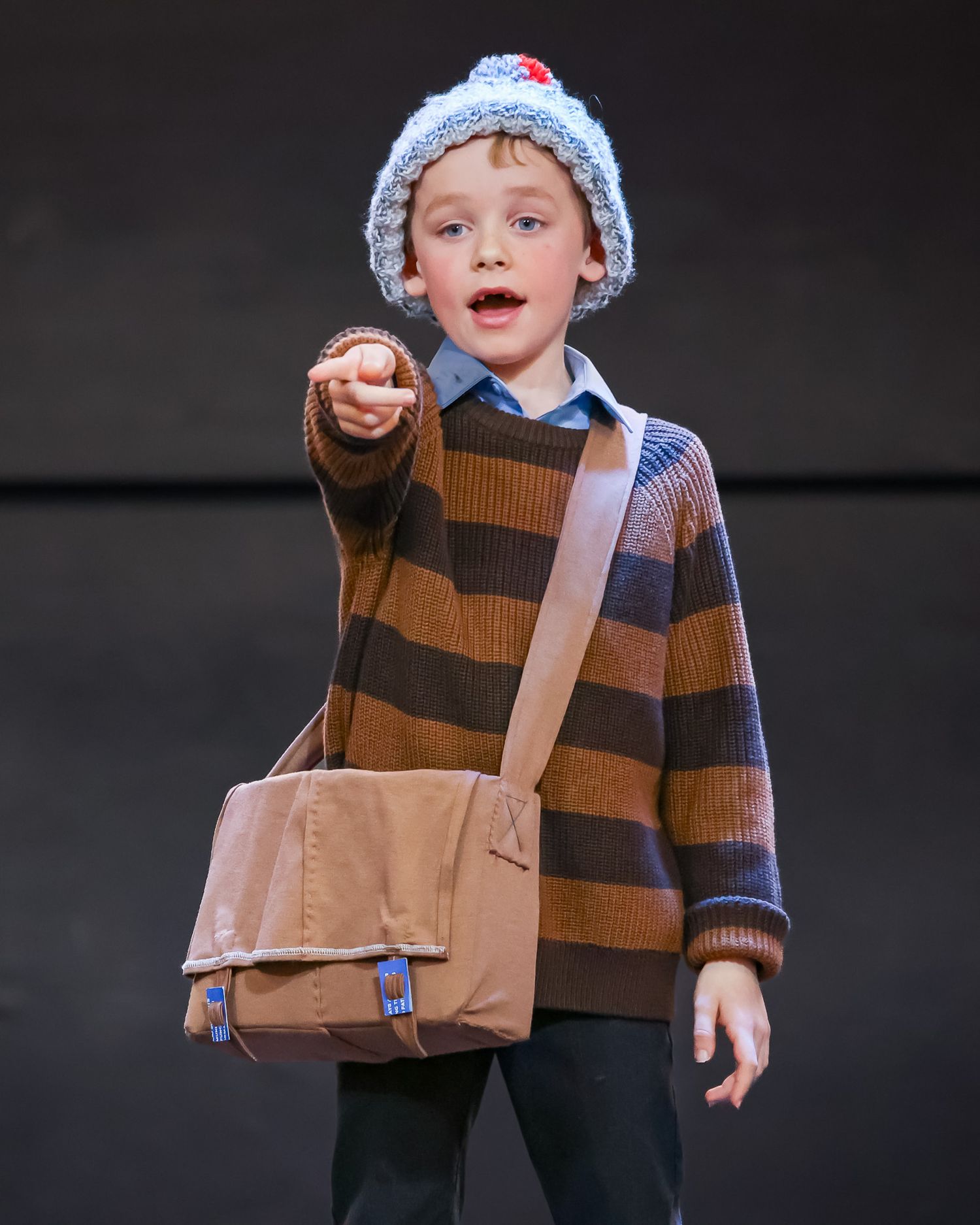 Young boy in striped sweater and knit hat pointing forward while carrying a tan messenger bag on stage.