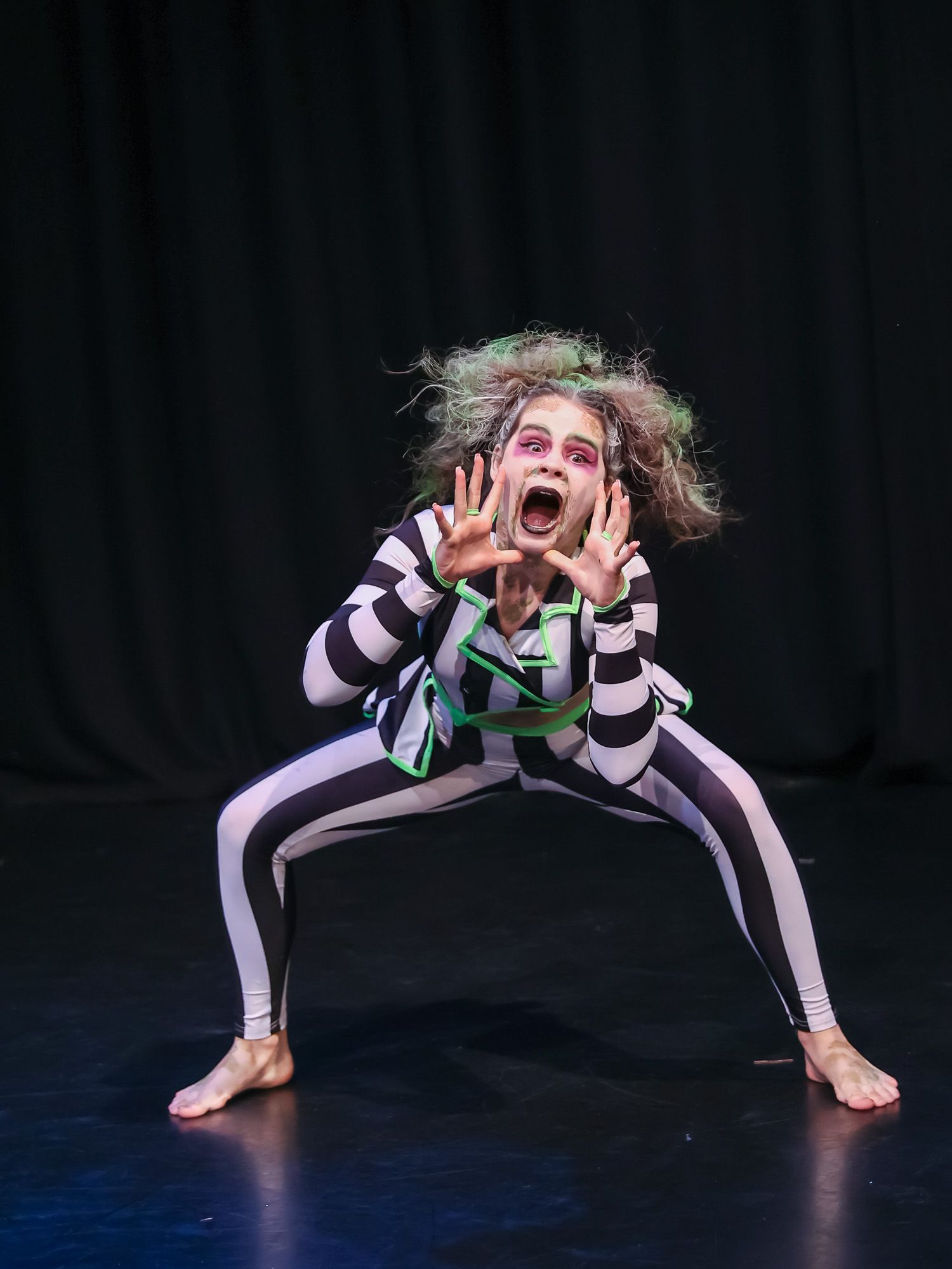 Child dancer in Beetlejuice black and white striped costume with green hair performs on stage.