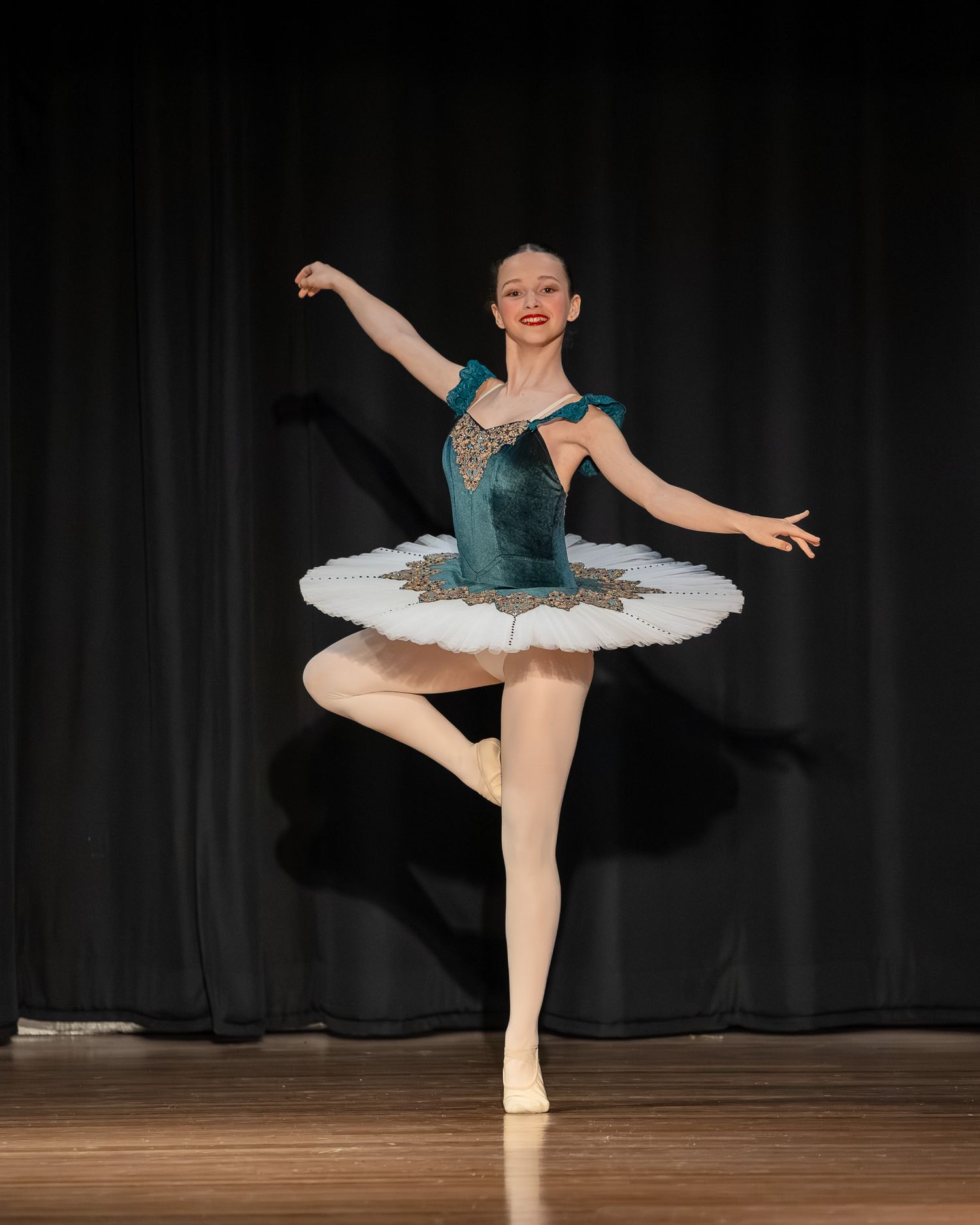 Young ballet dancer performs on stage in teal velvet tutu, executing a graceful one-legged pose under spotlight.