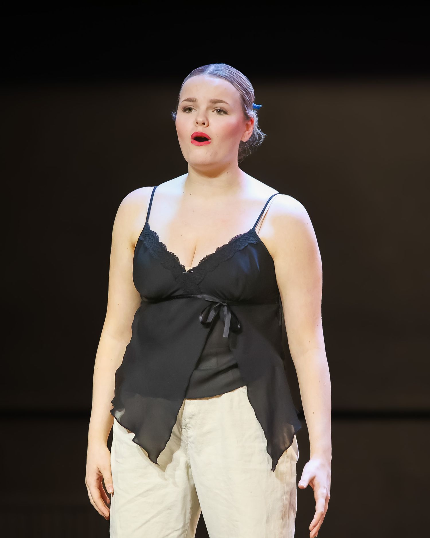 Young woman in black lace camisole and cream pants performing on stage with red lipstick.