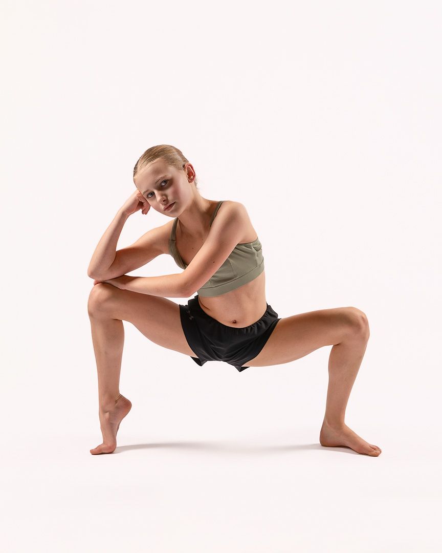 Dancer in athletic wear strikes a contemplative pose in wide stance against white backdrop.