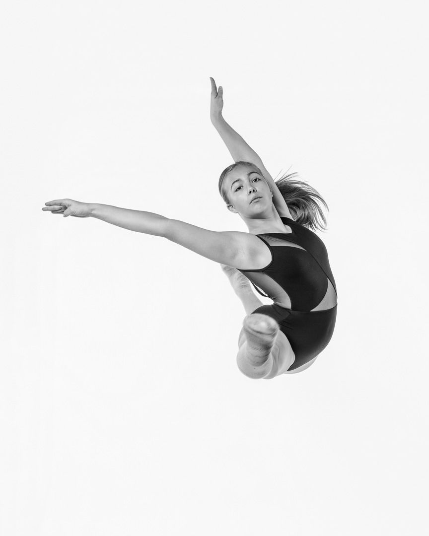 Dancer executes graceful leap with extended arms and legs in black leotard against white backdrop.