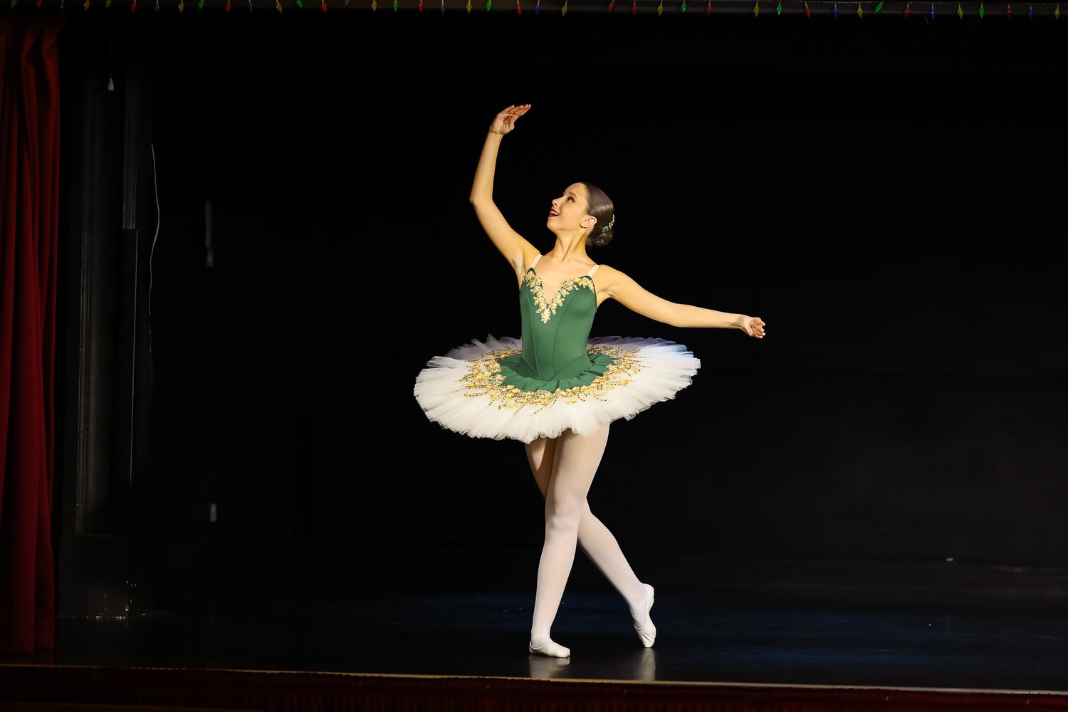 A ballerina in a green and white tutu performs an elegant pose on stage under bright lights.