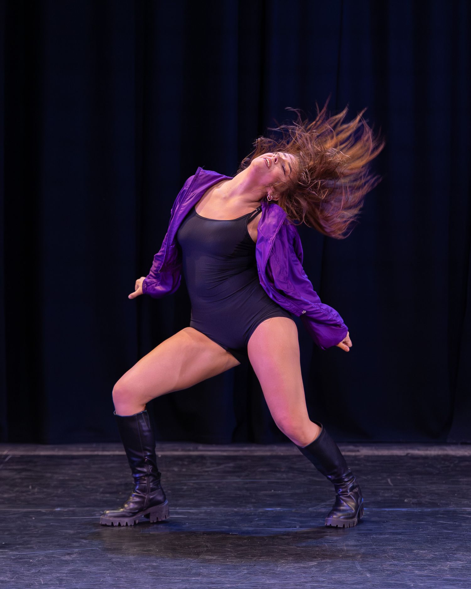 Female dancer in black leotard and purple jacket performs dramatic hair flip on stage wearing black boots.