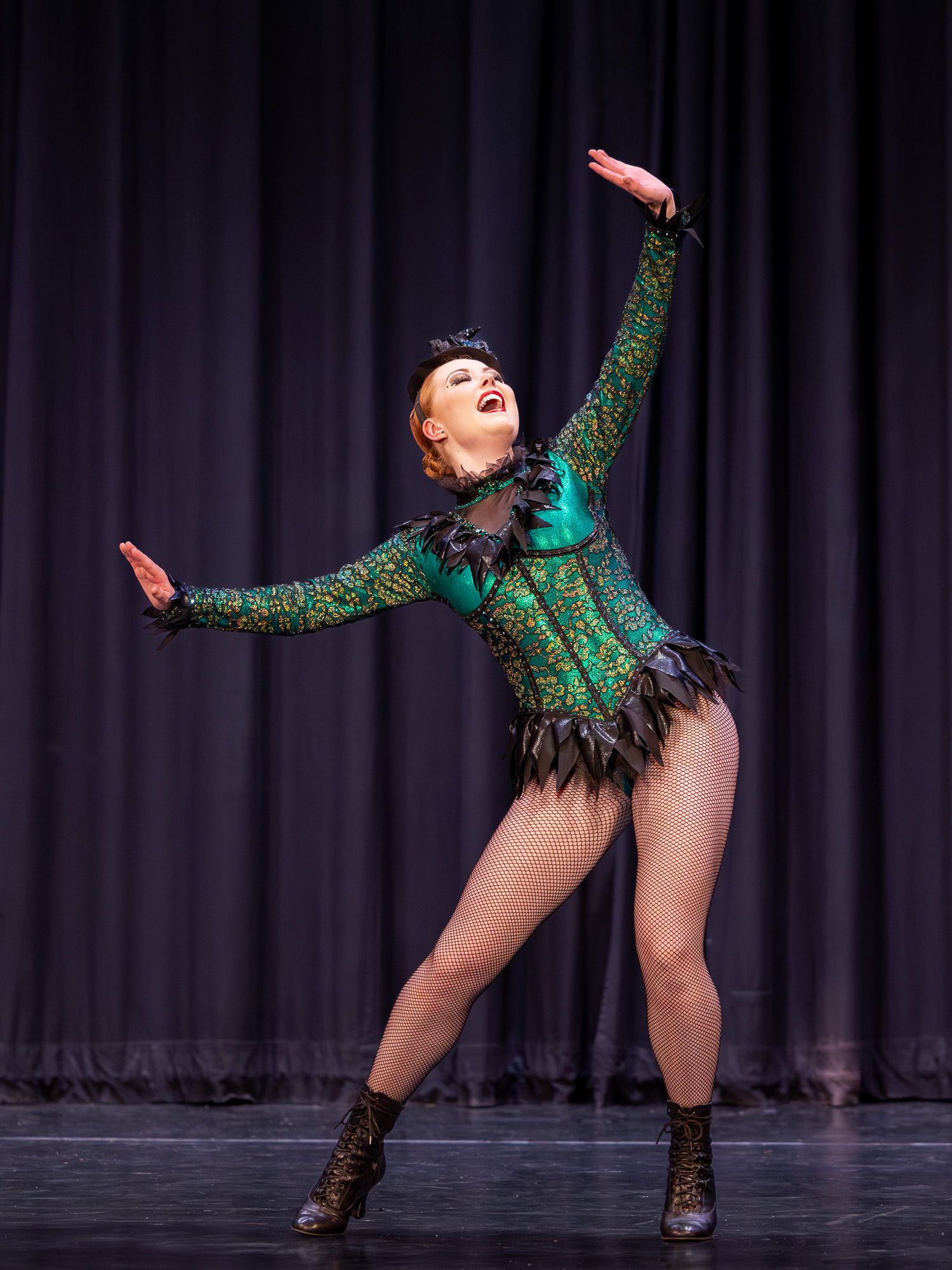 A dancer in a green sequined costume with black feathers performs an expressive pose on stage under bright lights.