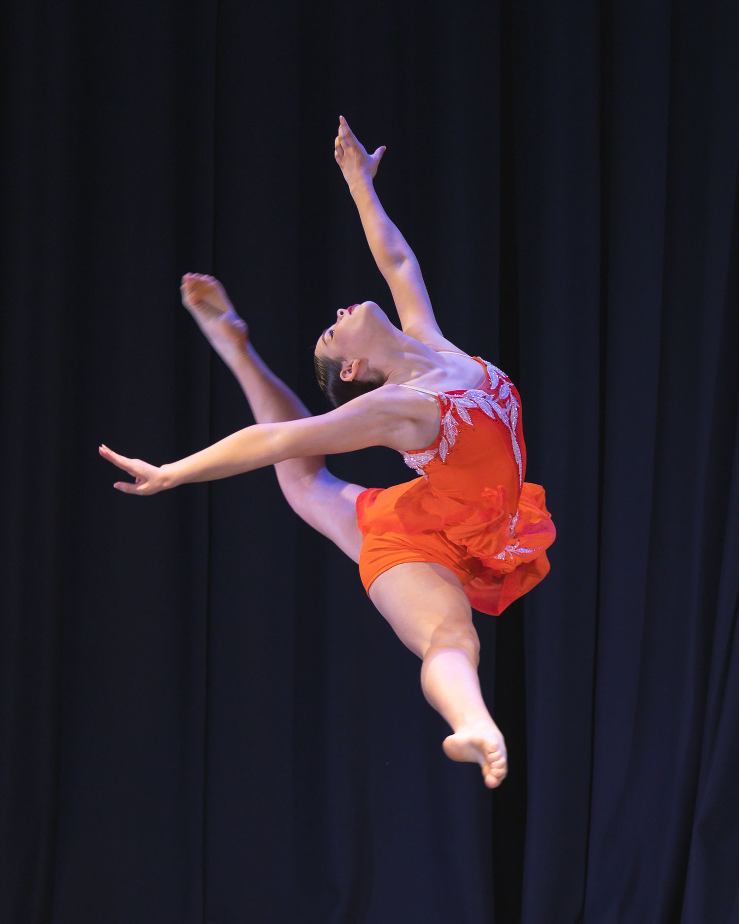 Dancer in orange costume performs graceful aerial move during stage performance.