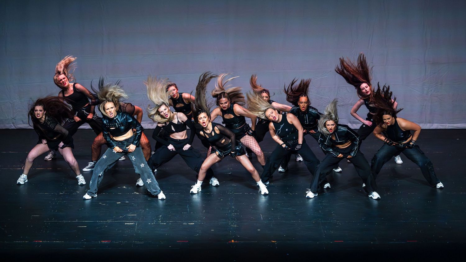 A large hip-hop dance crew performs energetically on stage, with dancers flipping their hair in synchronized choreography.