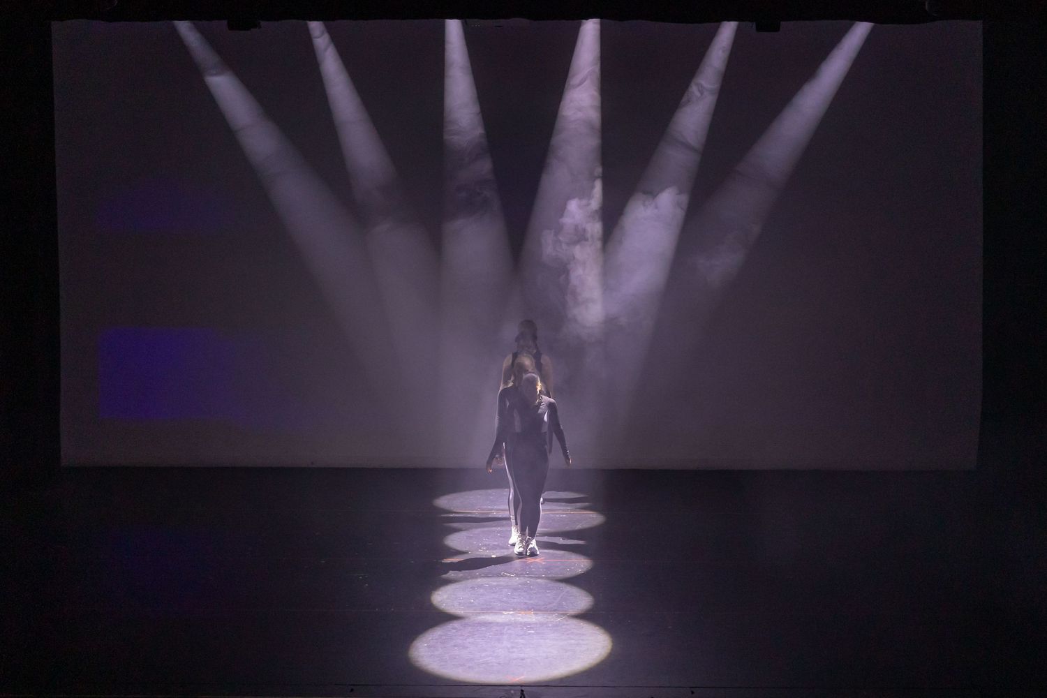 A lone performer walks on a dark stage illuminated by dramatic spotlights and haze during a live show.