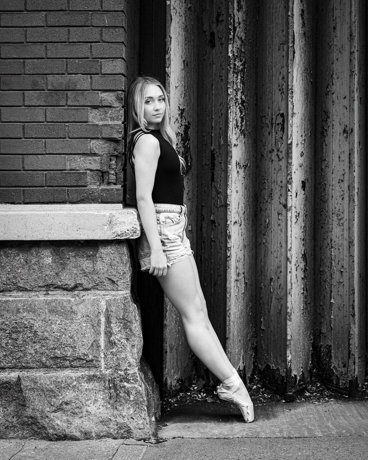 Black and white photo of a young female ballet dancer in pointe shoes posing gracefully against a rustic brick wall.