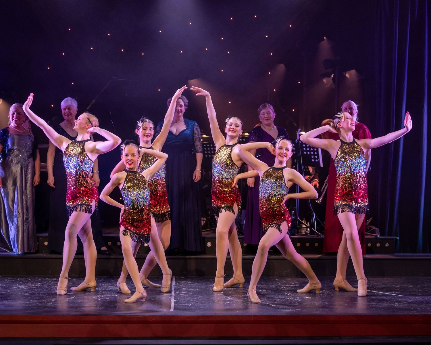 Young dancers in sequined costumes perform on a lit stage during a dance recital or show.