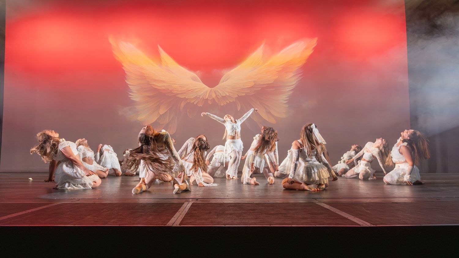 Dancers in white costumes perform on stage with a glowing angel wings backdrop in a dramatic dance recital.