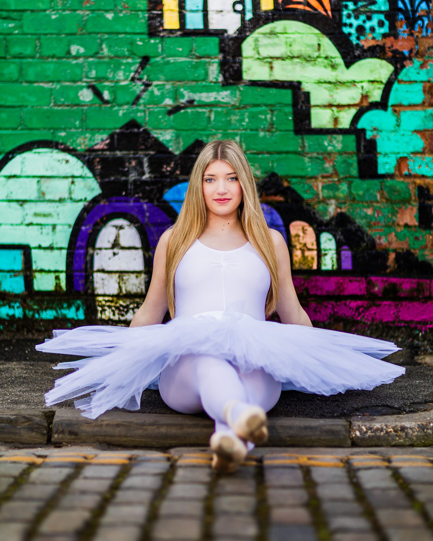 Young ballerina in white tutu and leotard sits gracefully on pavement before vibrant graffiti wall.