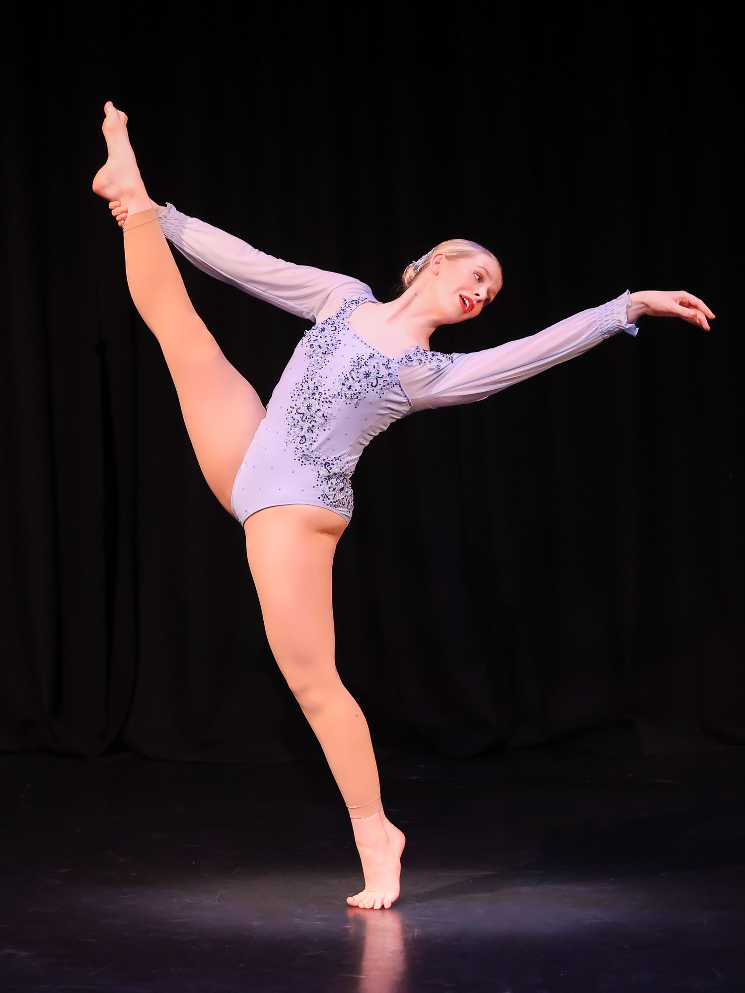 A young ballet dancer performs an arabesque in a blue leotard on a dark stage.
