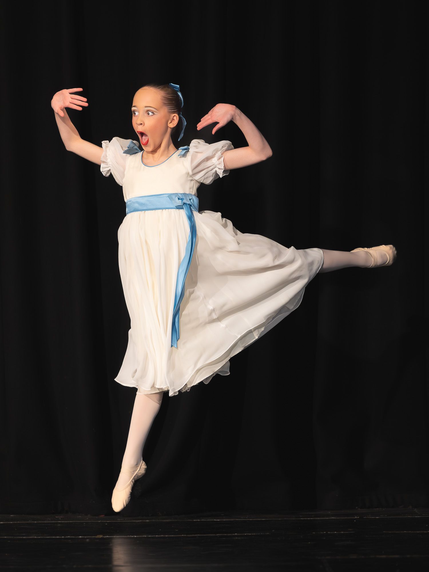 Young ballet dancer in white dress with blue sash leaps gracefully on stage during a performance.