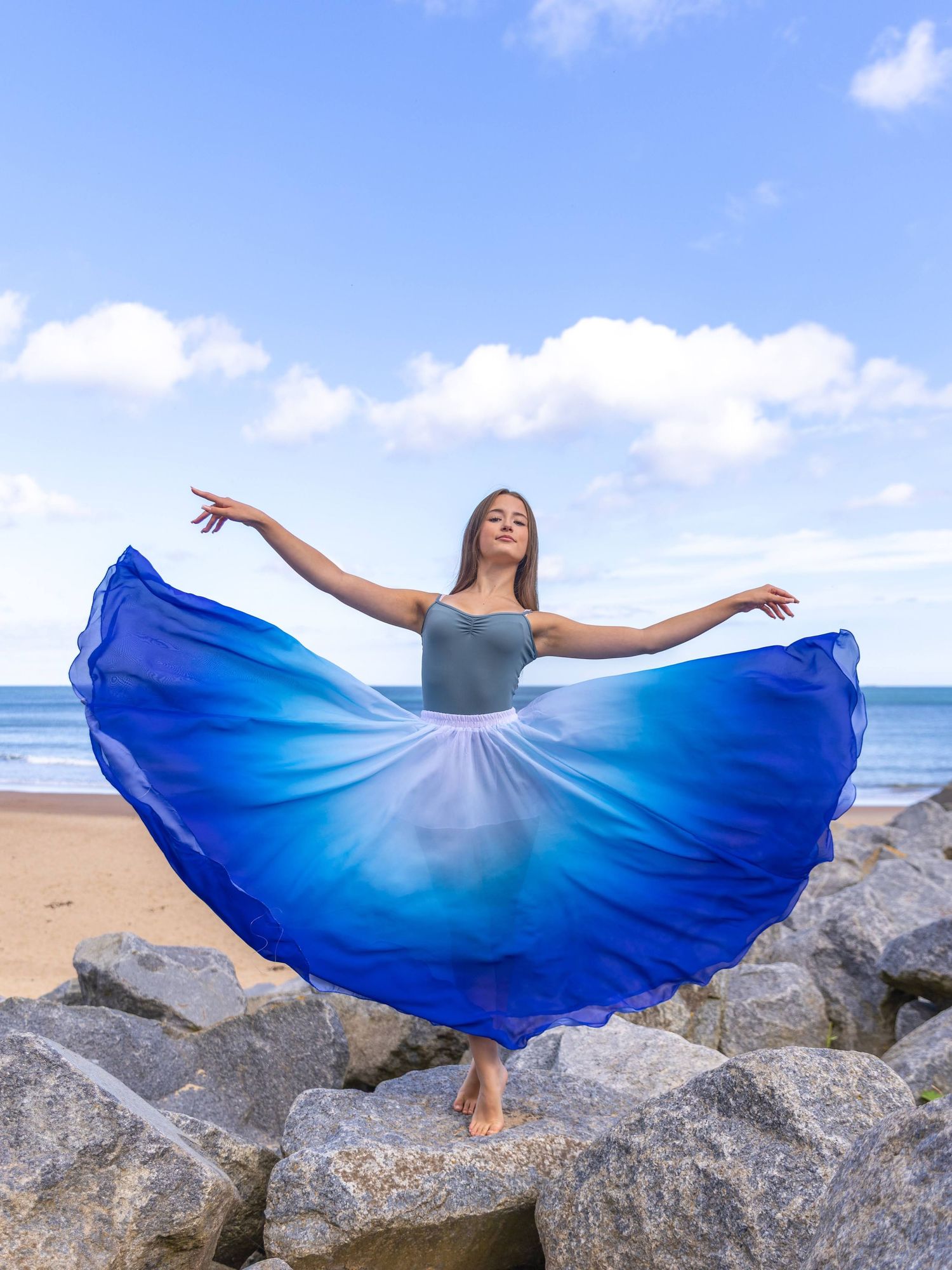 A dancer in a flowing blue ombre dress poses gracefully on coastal rocks with ocean and sky in the background.