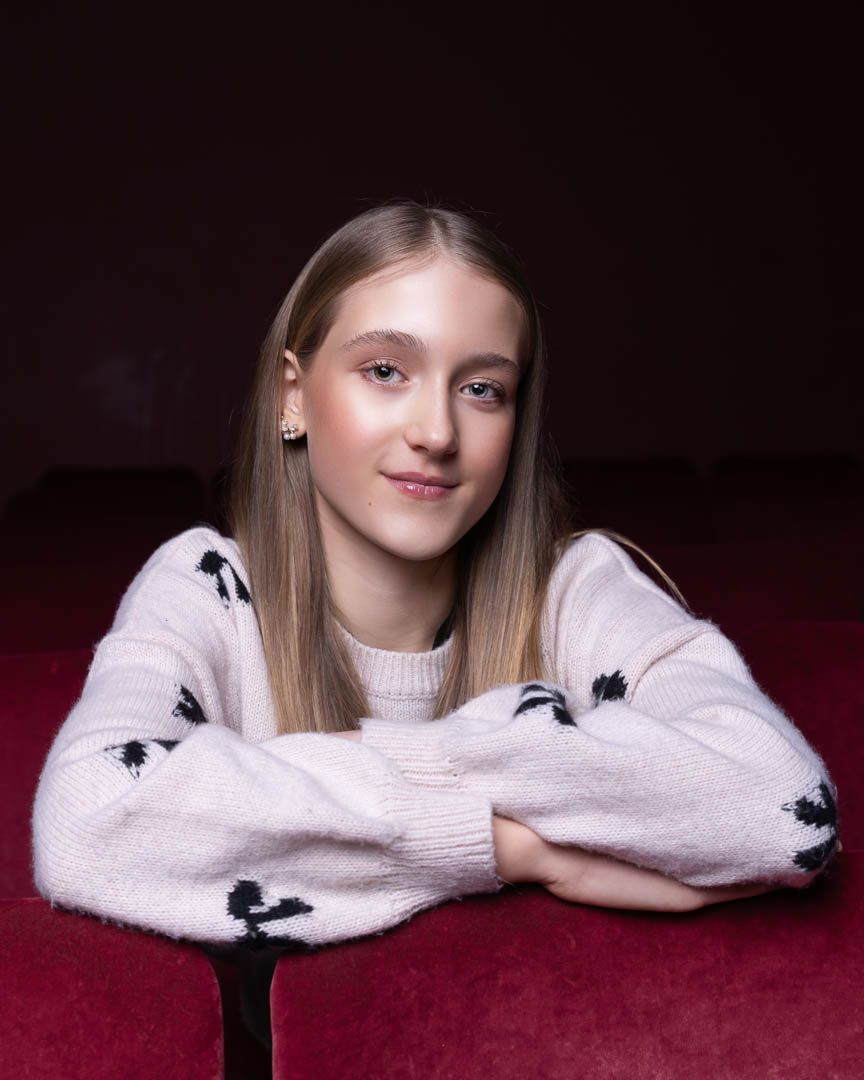 Young blonde girl in white patterned sweater poses with arms crossed, smiling against dark red background.